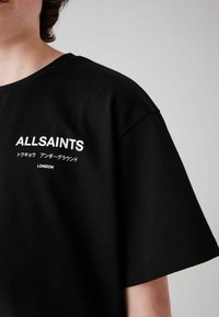 Černé tričko s krátkým rukávem, vyrobené z bavlny. Na přední straně je vytištěno jméno značky "ALLSAINTS" a text ve třech jazycích.