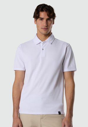 Junger Mann mit dunklen Haaren, der ein schlichtes weißes Poloshirt mit kurzen Ärmeln und beige Hosen trägt, vor einem neutralen Hintergrund stehend.