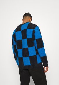 Sininen ja musta ruudullinen neule, pörröinen rakenne, rento malli, pyöreä kaula-aukko, ribbaatut hihat, näkyvät saumat, yhdistetty tummiin housuihin.