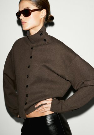 Donna che indossa maglione marrone scuro con collo alto e bottoni, occhiali da sole neri e pantaloni in pelle neri con mano sul fianco su sfondo neutro.