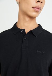 Crna pamučna polo majica s klasičnim ovratnikom, placketom sa dva gumba i urezanim Levi's logotipom na prsima. Glatka tekstura.