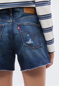 Γυναίκα που φοράει ψηλόμεσο μπλε τζιν σο ρτς με ξεφτισμένες άκρες και ριγέ μακρυμάνικο πουκάμισο, εμφανιζόμενη από πίσω.