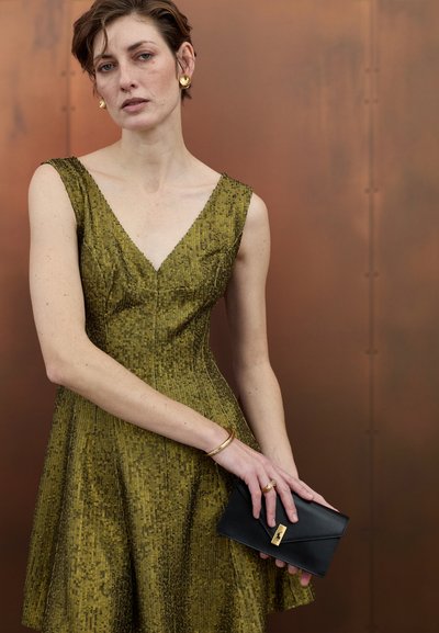 Vestido ajustado de color verde oliva con escote en V y tejido texturizado. La modelo sostiene un bolso negro de mano con un detalle en dorado. Unos sencillos complementos dorados completan el conjunto.