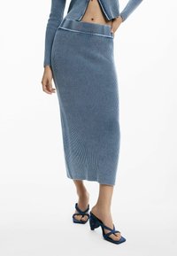 Vrouw in een ribbelige blauwe gebreide midi rok met een bijpassende cropped cardigan en blauwe hoge sandalen met kruislingsel straps.