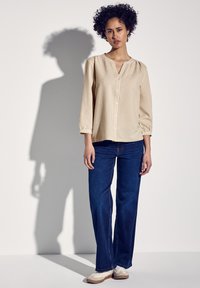 Camicia a quadri color beige chiaro con un taglio rilassato, scollatura a buco chiave e maniche a sbuffo, abbinata a jeans larghi di colore blu scuro e sneakers bianche.