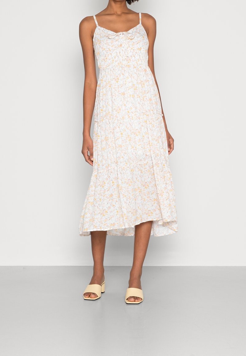 Vestido midi blanco con motivos florales y acentos amarillos, tirantes finos ajustables y un cuerpo ceñido, que fluye hacia un dobladillo en capas. Llevado con sandalias beige.