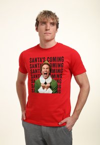 Červené bavlněné tričko s potiskem postavy v zelené barvě s bílým límcem a černým nápisem "SANTA'S COMING".