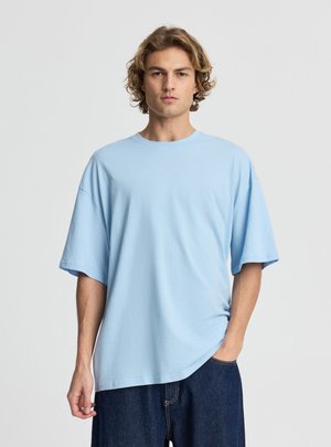 Giovane uomo con capelli biondi ondulati che indossa una maglietta azzurra larga e jeans scuri con una mano in tasca su sfondo semplice.