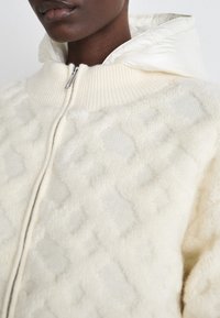 Krämfärgad zip-up jacka med kviltad textur, stickad krage och glänsande vit huva innanför. Mjuk tyg med upphöjd diamantmönster.