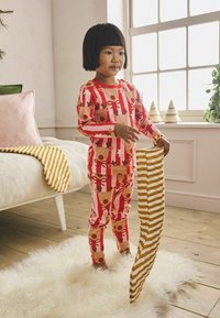 Punane ja valge triibuline laste pyjama, millel on põhjapõdra muster. Laps hoiab triibulist koorvalget ja kuldset tekk. Puitpõrand on nähtav.