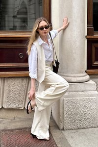 Balts piegrieztais vestes uz baltas blūzes, apvienots ar krēmkrāsas platām biksēm un melniem jostainiem sandaliem. Brūna plecu soma izceļ šo izskatu.