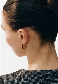Guldörhänge i form av en rund ring på en modells öra, med en slät, böjd design. Bakgrunden är minimal, vilket framhäver accessoarens glans och form.