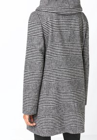 Henkilö, jolla on mustavalkoinen, koiranhammaskuviollinen takki, jossa on huppu, esitettynä takaa puoliväliin reittä.