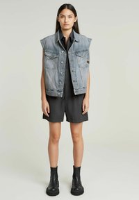 Gilet di jeans in lavaggio chiaro con grandi tasche frontali, indossato sopra una tuta nera, abbinato a stivaletti neri. Texture liscia, design oversize.