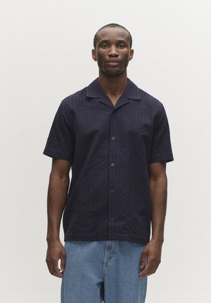 Muž oblečený v tmavomodrej krátkorukávovej košeli na gombíky a svetlomodrých džínsach, stojaci pred jednoliatym bielym pozadím.