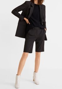 Ensemble blazer et short à fines rayures noires, associé à un top noir et des bottines crème. Tissu lisse, coupe ajustée et design structuré.
