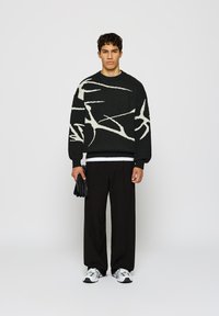 Joven de pie, vistiendo un suéter abstracto en blanco y negro, pantalones anchos negros, zapatillas blancas, sosteniendo guantes negros en la mano izquierda.