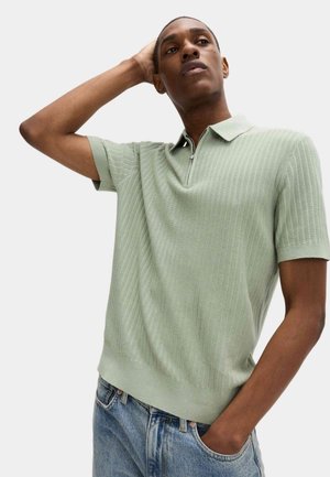 Uomo che indossa una polo a maniche corte a coste verde chiaro con cerniera, in posa con una mano sulla testa e l'altra in tasca, su sfondo uniforme.