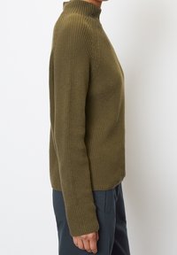 Olivegrüner Strickpullover mit hohem Kragen, gerippter Textur, langen Ärmeln und tailliertem Schnitt, kombiniert mit marineblauen Hosen.