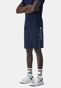 Pantalones cortos deportivos de color navy con rayas laterales y logo, combinados con zapatillas blancas y calcetines con rayas negras. Los pantalones cortos tienen una cintura elástica.