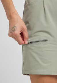 Grüner, leichter Stoffrock mit einer seitlichen Reißverschlusstasche, der eine glatte Textur und klare Linien aufweist. Hand dezent in der Nähe des Reißverschlusses platziert.