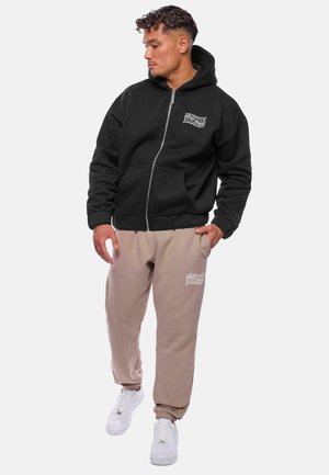 Sort zip-up hoodie med en kængurulomme, parret med beige joggers med et broderet logo, båret med hvide sneakers.