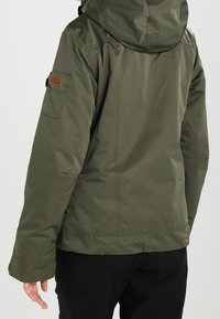 Veste kaki verte avec capuche, en tissu texturé, avec poche latérale et détails de couture visibles. Design épuré avec une silhouette ajustée.