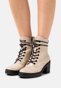 Bottines en cuir verni beige avec lacets noirs, bout arrondi et talon épais. Dotées d'une fermeture éclair latérale et d'une semelle texturée pour une meilleure adhérence.