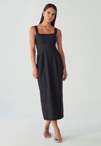 Ženska stoji v brezrokavni črni midi obleki s kvadratnim izrezom in bež sandali s trakovi pred enobarvno svetlo ozadje.