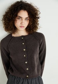 Cardigan texturizado castanho com um decote redondo e seis botões dourados à frente; emparelhado com uma saia riscada, possui mangas longas.