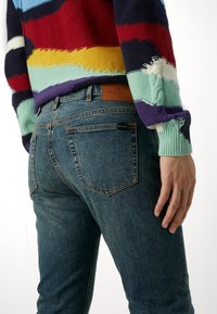 Jeans de mezclilla azul con ajuste slim, con bolsillos traseros y una etiqueta de cuero marrón con la inscripción "PS Paul Smith." Suéter de punto texturizado con rayas vibrantes.