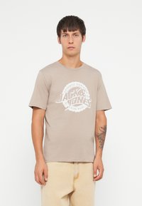 Hvit beige herret-skjorte laget av bomull, med et sentralt hvitt grafisk logo. Korte ermer og rund hals.
