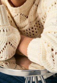 Person som bär en krämfärgad stickad tröja med ett utskärningsmönster, vilar armarna på en glänsande metallyta, med synliga blåjeans.