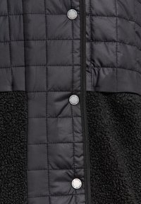 Musta tikattu toppatakki, jossa on pehmeä fleece-alapala, painonapilliset sulkimet ja materiaalien vastakkaiset tekstuurit.