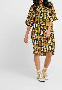 Zelené šaty s vzorem a béžovými a bílými abstraktními tvary, střední délky, volného střihu; doplněné béžovými vysokými teniskami a bílými ponožkami.