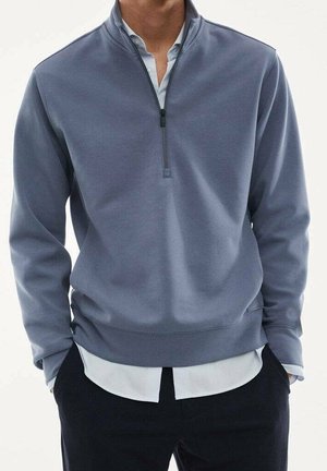 Hombre con un suéter de cremallera hasta un cuarto en azul sobre una camisa blanca y pantalones negros, con las manos en los bolsillos, frente a un fondo llano.