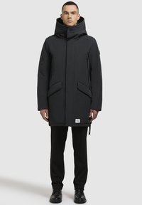 Parka neagră, izolată, cu guler înalt, două buzunare frontale, buzunare laterale cu fermoar și o croială dreaptă. Textură netedă și design minimalist.
