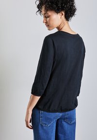 Schwarzes, leichtes Oberteil mit 3/4-Ärmeln, entspannter Passform und runder Ausschnitt, kombiniert mit blauen Jeans. Glatte Textur, minimalistisches Design.