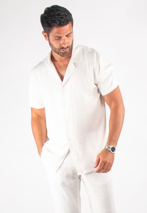 Chemise en lin blanc à manches courtes avec col, présentant un tissage texturé et une fermeture à boutons sur le devant. Associée à un pantalon en lin assorti.