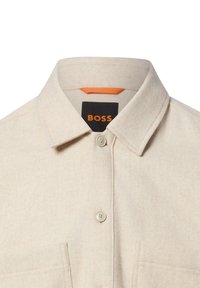 Beżowa flanelowa koszula z kołnierzykiem na guziki, dwoma przednimi kieszeniami oraz czarną metką z pomarańczowym napisem wskazującym markę "BOSS".