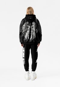 Chaqueta bomber negra con diseño de alas plateadas en la parte posterior, combinada con pantalones negros con un detalle rasgado y etiqueta blanca, y botas blancas.