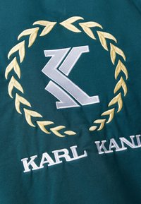 Tessuto verde scuro con una ghirlanda di foglie dorate ricamata e il logo "K" in bianco, con il testo "KARL KANI" sotto in cucitura bianca.