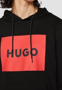 Sweat à capuche noir avec un grand graphique rectangulaire rouge portant le texte "HUGO" en noir. Comprend des cordons de serrage et un tissu texturé.