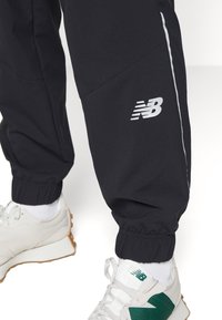 Black jogger pants with elastic cuffs, featuring a white side stripe and a grey New Balance logo on the right thigh.