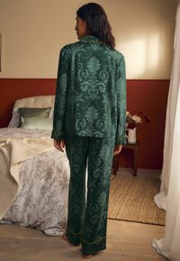 Zelený pyžamový set so vzorom obsahujúcim kvetinové motívy, košeľa s golierom a dlhými rukávmi, a zladené nohavice so zlatými akcentmi.