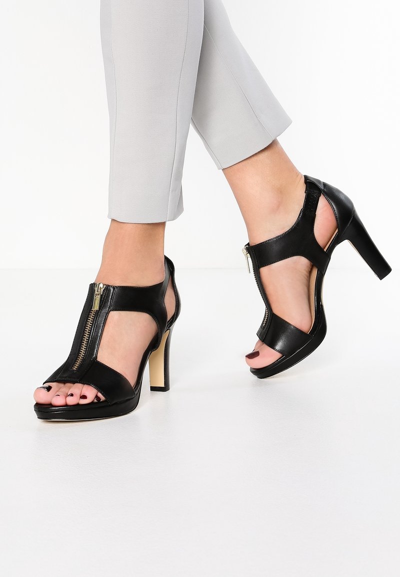 Sandales à talons hauts en cuir noir avec un bout ouvert, un design découpé et une fermeture éclair à l'avant. Les talons sont épais, contrastant avec une finition lisse.