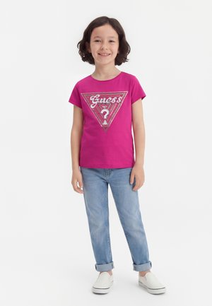 Παιδί με κοντά σκούρα μαλλιά, που φοράει φωτεινή ροζ μπλούζα Guess, ανοιχτό μπλε τζιν με γυρισμένα μπατζάκια και λευκά παπούτσια τύπου slip-on, χαμογελάει και στέκεται όρθιο.