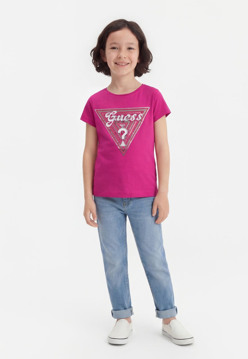 Παιδί με κοντά σκούρα μαλλιά, που φοράει φωτεινή ροζ μπλούζα Guess, ανοιχτό μπλε τζιν με γυρισμένα μπατζάκια και λευκά παπούτσια τύπου slip-on, χαμογελάει και στέκεται όρθιο.