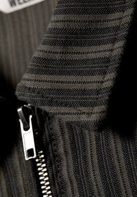 Zwart en grijs gestreepte stof met een gestructureerd oppervlak, voorzien van een zilveren rits en een kraagaccent. Close-up weergave.