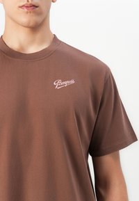 Close-up of a person wearing a brown t-shirt with a small pink "Pompeii" logo on the left chest against a plain white background.